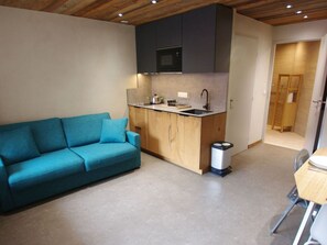 Ein kompakter Wohnraum mit Küchenzeile, einer Couch, einem Mülleimer und einer Tür zu einem weiteren Zimmer.