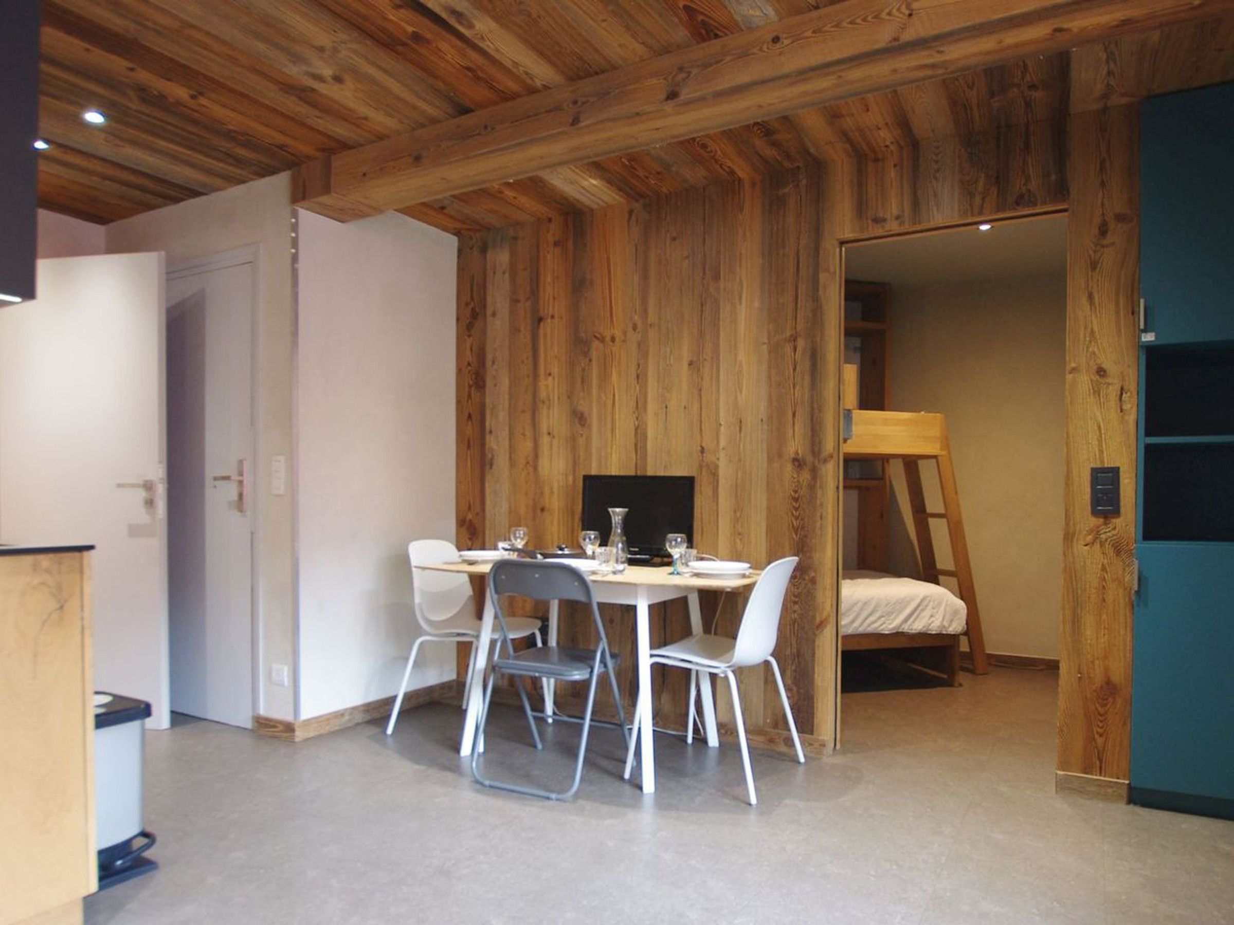 A room with a wooden ceiling, a dining table with chairs, a bed, and a blue cabinet.