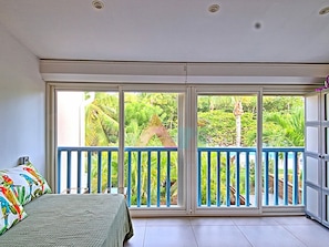 A room with a bed, a balcony with a blue railing, and a view of greenery outside. - Duplex house with air conditioning - 6 guests (Saint-Martin)