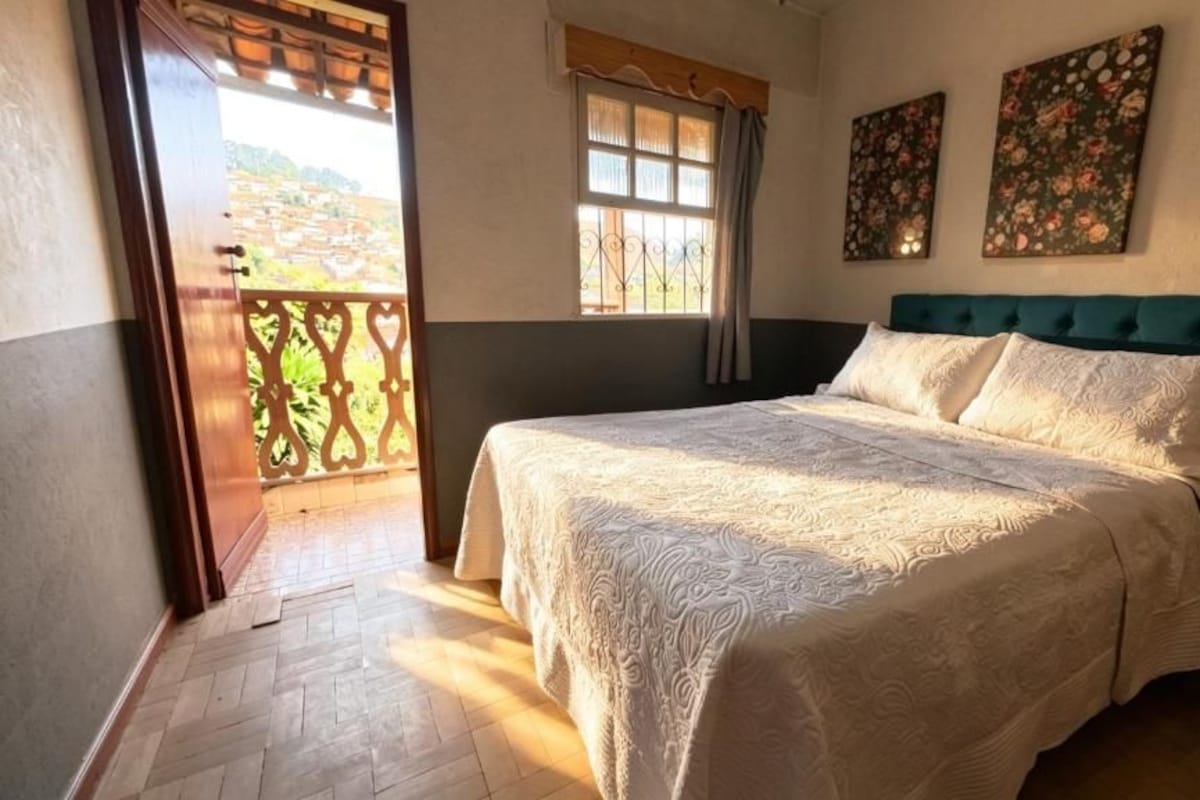 A bedroom with a bed, a window, and a view of a balcony.