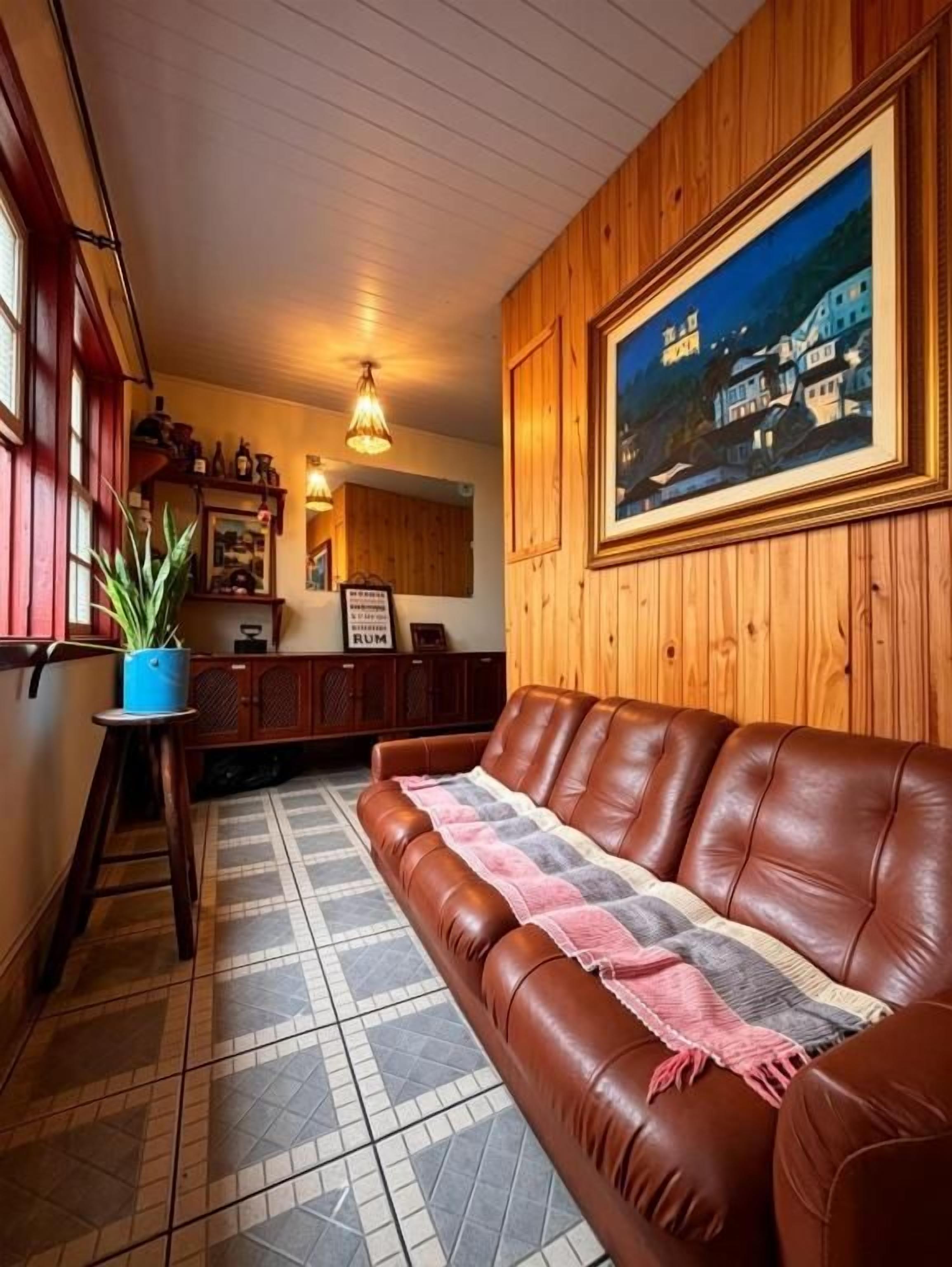 Ein Zimmer mit einem braunen Ledersofa, einem gerahmten Gemälde, einem Blumentopf und einem Fliesenboden.