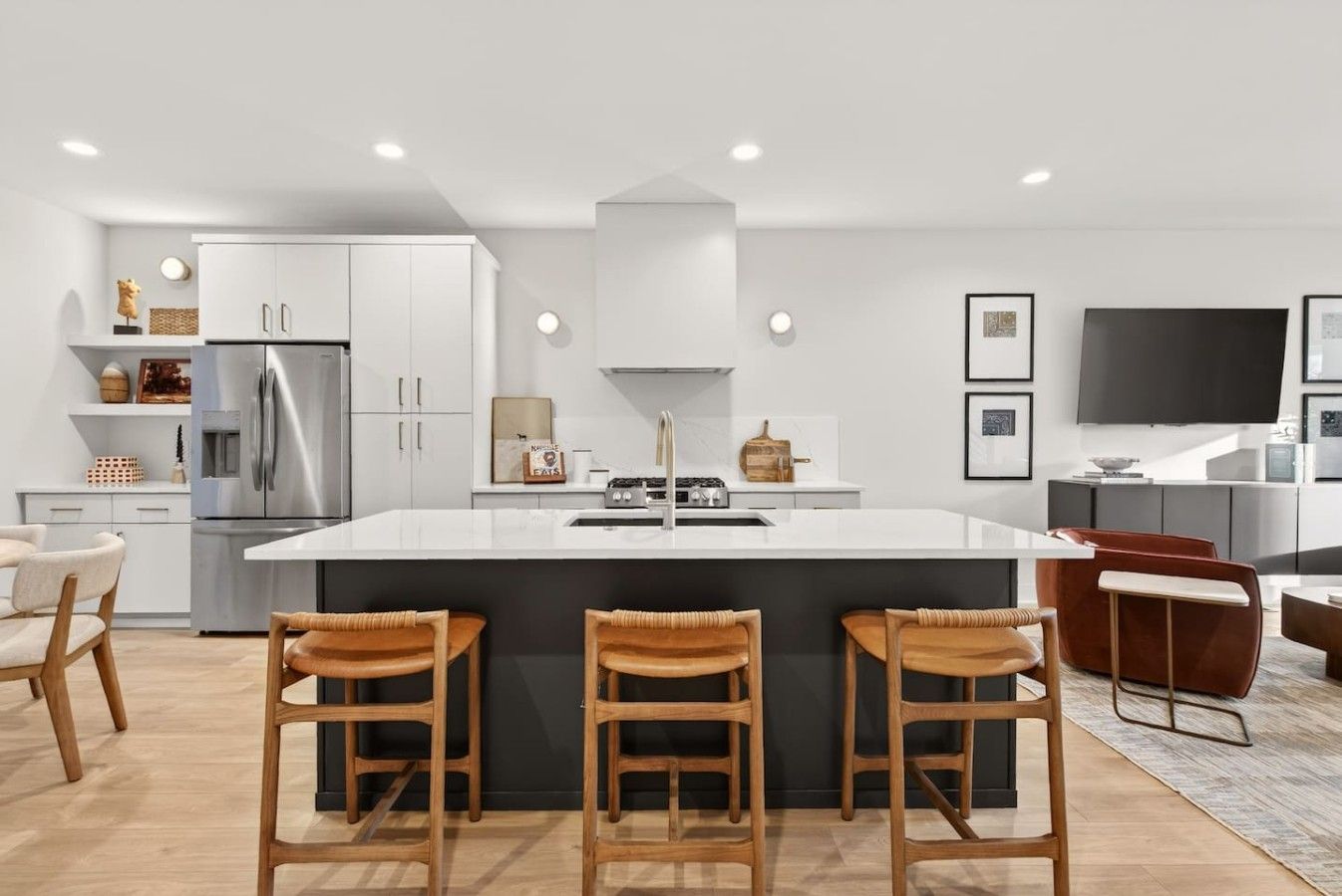 A modern kitchen with a central island, bar stools, a refrigerator, and a flat-screen TV.