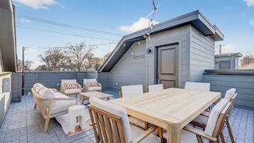 A covered patio with a wooden table and chairs, a small building, and a clear sky.