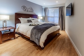 A modern bedroom with a wooden bed, bedside tables, a flat-screen TV, and a window with curtains. - Ski-in/Ski-out 1Bdrm Luxury Condo, Slps 4, 1st fl, The Aspens, Pool & hot tubs (Whistler)