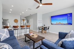 Ein modernes Wohnzimmer mit Fernseher, der die Öffnungszeiten von Disney Springs zeigt, einem Essbereich mit rundem Tisch und einer Küche im Hintergrund.