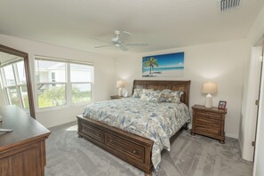 Une chambre à coucher comprenant un lit, deux tables de chevet, un ventilateur de plafond, une fenêtre et un tableau représentant une scène de plage.