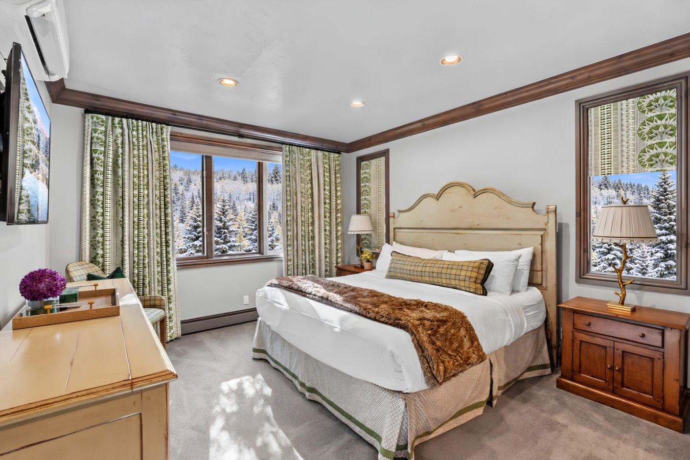 Un dormitorio con una cama grande, una mesita de noche, una lámpara y una ventana con vistas a árboles cubiertos de nieve.