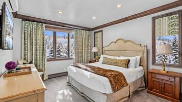 Un dormitorio con una cama grande, una mesita de noche, una lámpara y una ventana con vistas a árboles cubiertos de nieve.