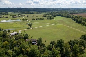 A large open field with a winding path, surrounded by trees and a few buildings. - Riverside house on a Duck River ranch: fishing lake, horses, covered deck (Normandy)