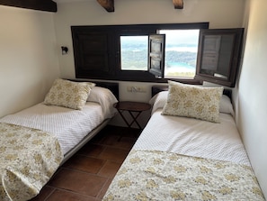 A small room with two beds, each with floral-patterned bedding, a small table, and a window with a view.