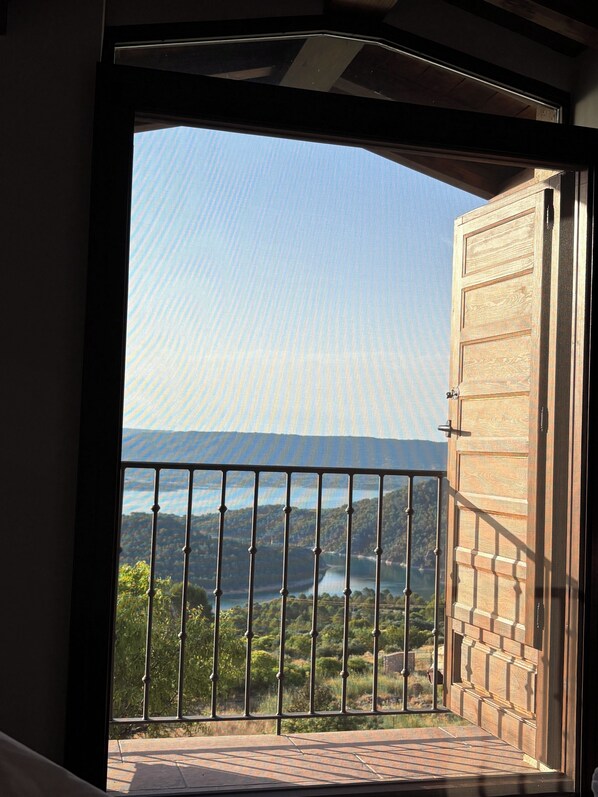 A view from a balcony with a metal railing, overlooking a landscape with trees and a body of water.