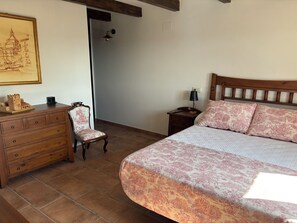 Un dormitorio con cama, cómoda, silla, mesita de noche, lámpara y un cuadro en la pared.