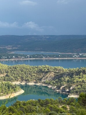 A coastal landscape with a clear view of a lake, dense greenery, and distant buildings. - Breathtaking Reservoir Views & Private Cave | 6-Bedroom Retreat Near Madrid (Alocén)