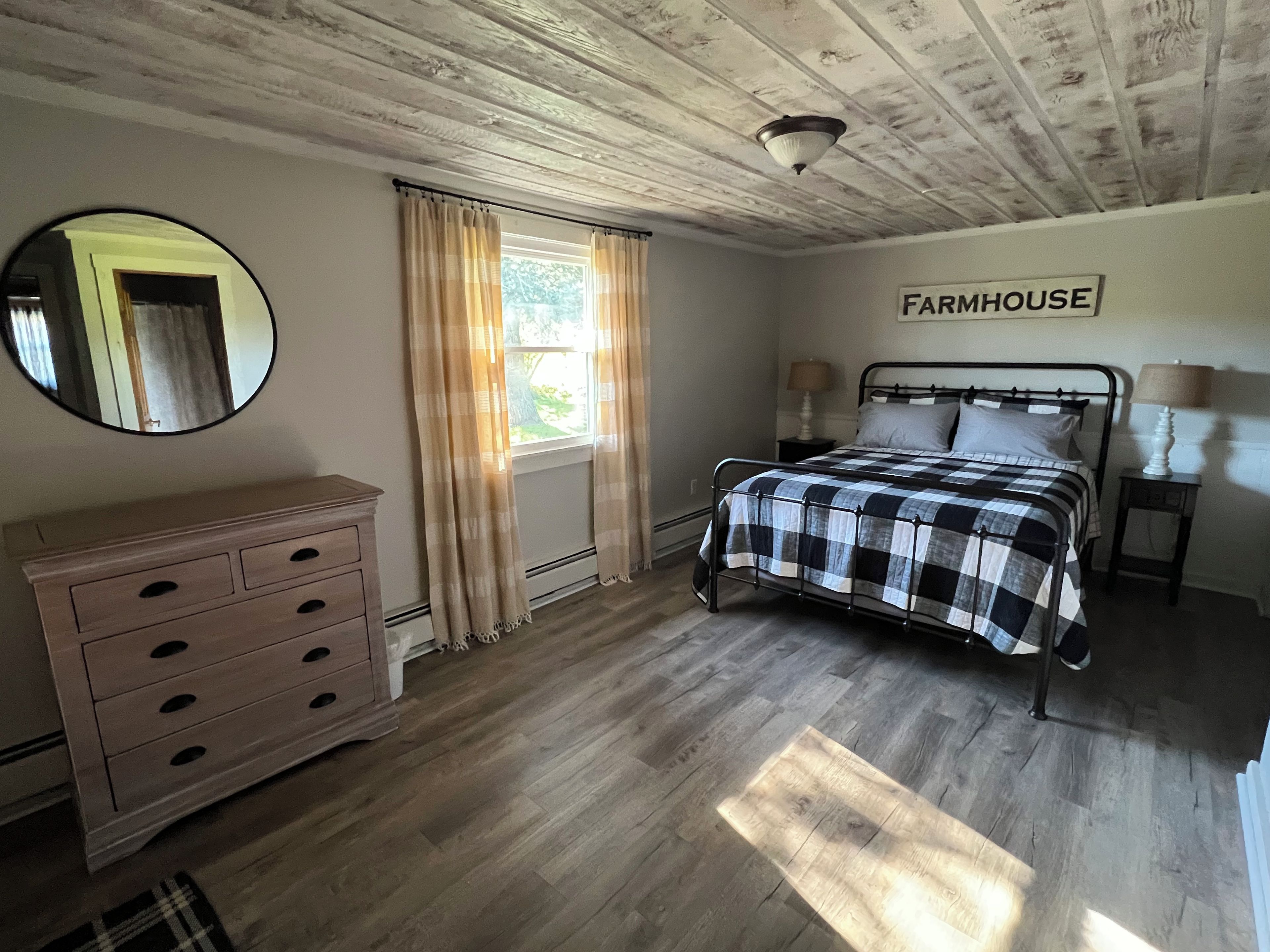 Ein Schlafzimmer mit Bett, Kommode, Spiegel, einem Fenster mit Vorhängen, Deckenventilator und einem Schild mit der Aufschrift 'FARMHOUSE'.