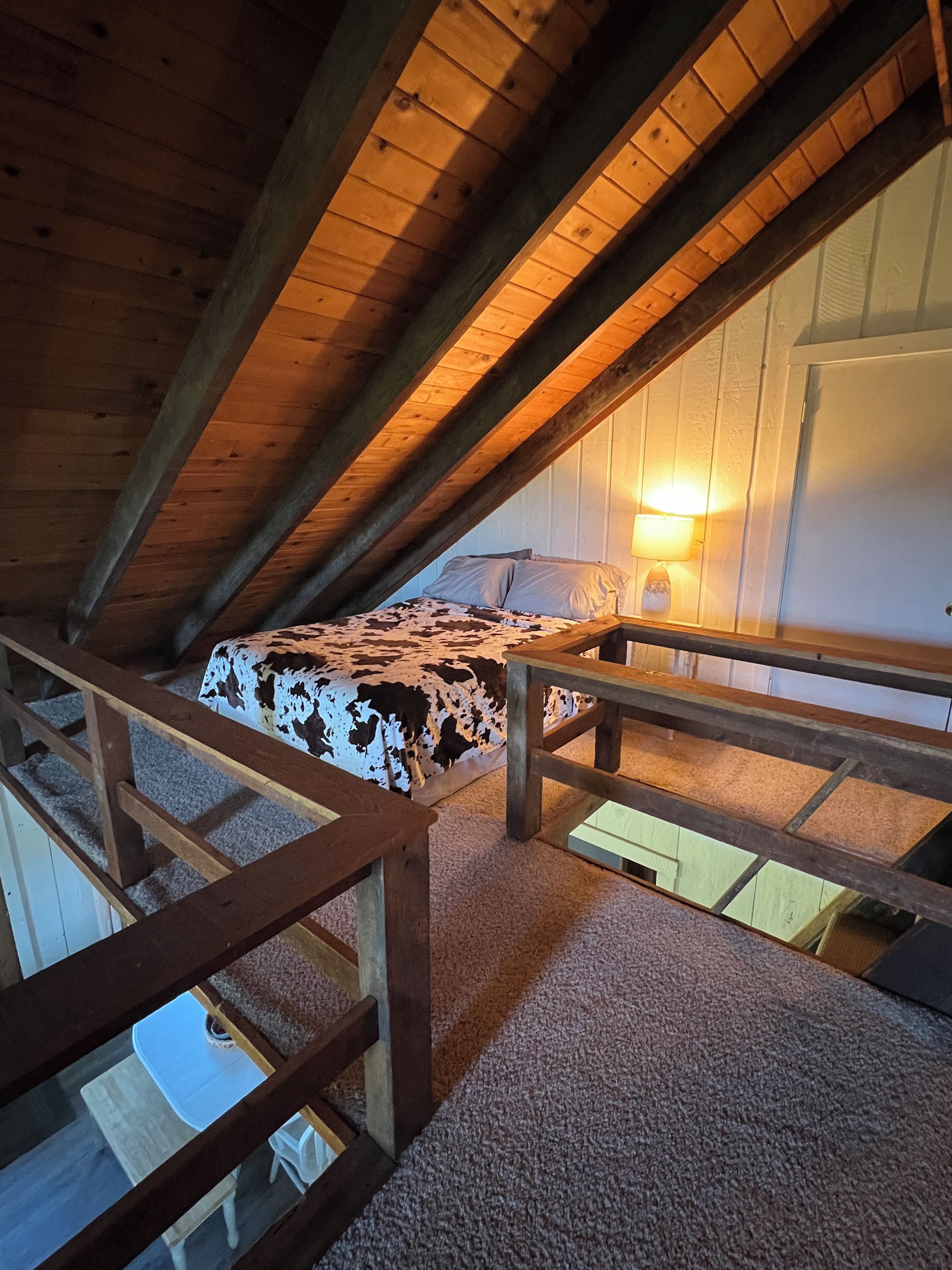 Un dormitorio en el ático con techo inclinado, una cama con colcha de vaca, una mesita de noche, una lámpara y vistas al exterior a través de una ventana.