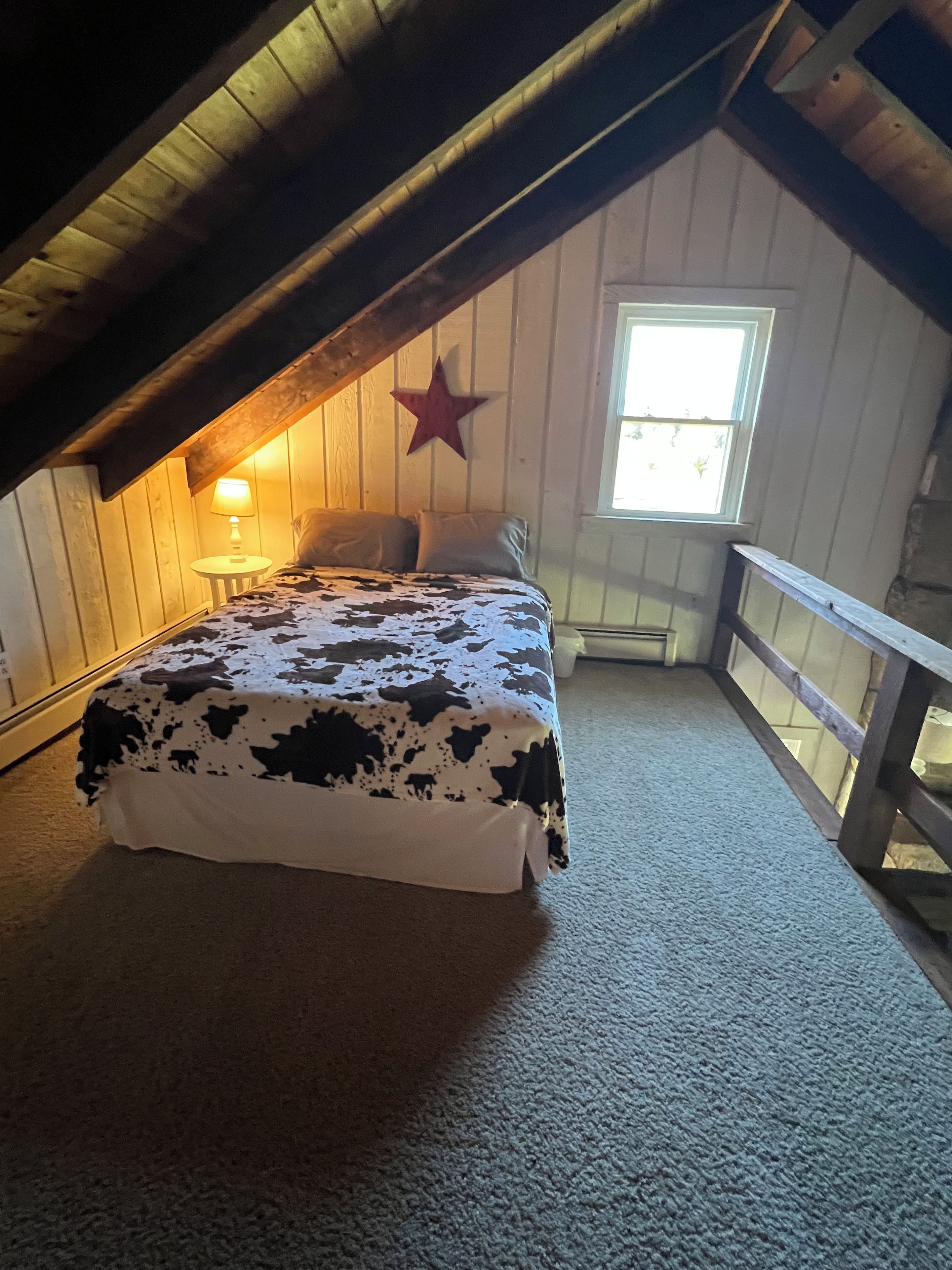 A bedroom with a sloped ceiling, a bed with a cow print bedspread, a star decoration, a window, and a nightstand with a lamp.