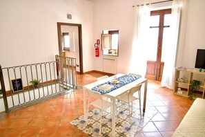A room with a tiled floor, a white table with a patterned tablecloth, a white chair, a wooden door, a window with curtains, a fire extinguisher, and a small shelf with a plant. - Sikelia Apartment—your retreat in the heart of Palermo. (Palermo)