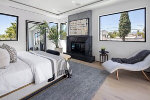 A modern bedroom with a large bed, a fireplace, a seating area with a chair, and large windows offering a view of the outdoors. - Casa Zenithia by Oraklus (Los Angeles)