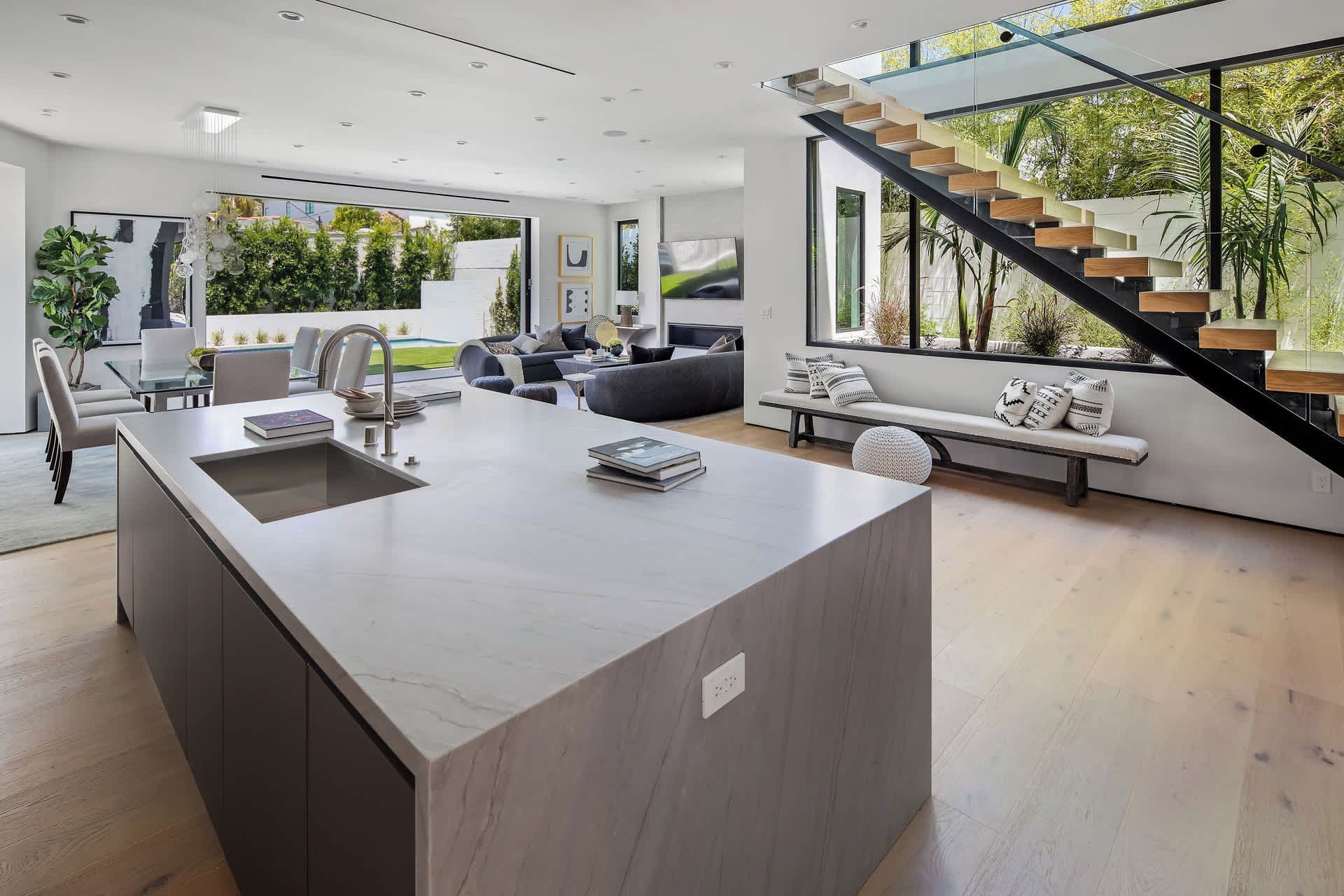 Une cuisine moderne dotée d’un îlot central, un coin repas avec une table en verre et un escalier menant à un balcon offrant une vue sur la verdure.