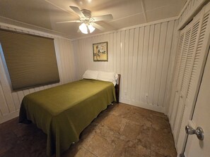 Ein Schlafzimmer mit einem Einzelbett, einem Deckenventilator, einem Fenster mit Jalousien, einer Tür mit Griff und einem Wandbild.