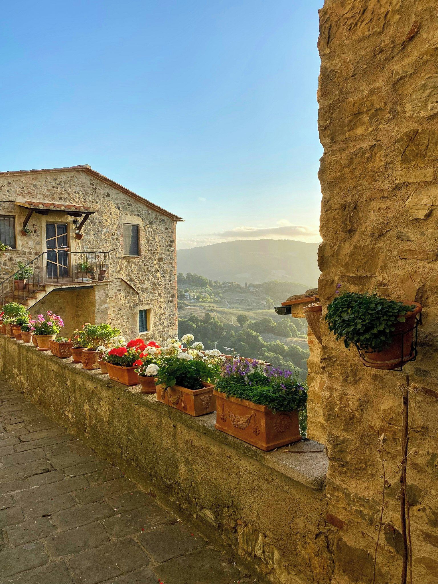 A stone building with a balcony, terracotta pots with flowers, and a view of a valley.