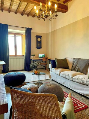 Ein gemütliches Wohnzimmer mit einer Couch, einem Esstisch, einer Chandelieber, einer Uhr und einem Laptop auf einem Schreibtisch.