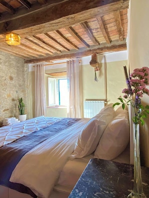 Un dormitorio con cama, mesita de noche, un florero con flores, ventana con cortinas y pared de piedra.