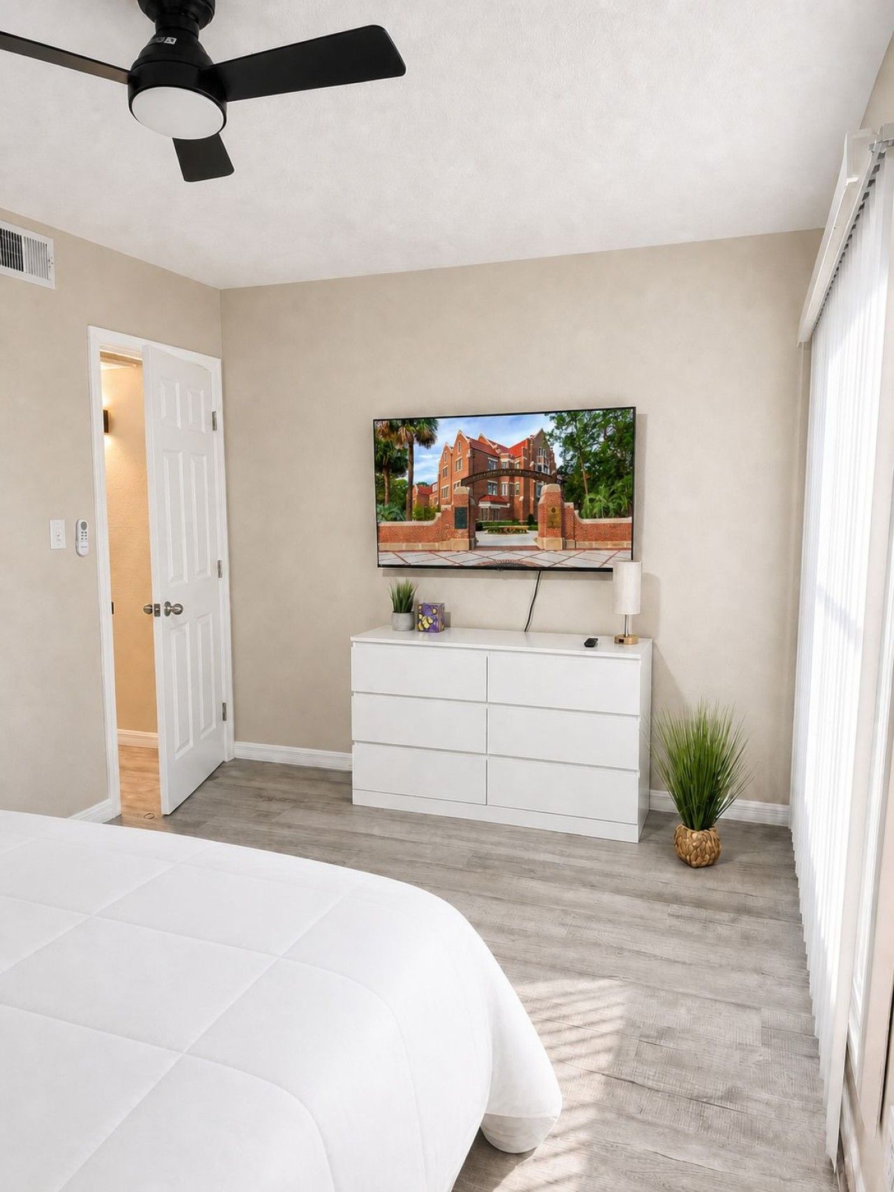 A bedroom with a bed, a ceiling fan, a white dresser, a television on the wall, and a door leading to another room.