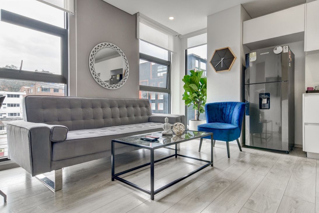 Una sala moderna con un sofá gris, una mesa de centro de cristal, un sillón azul, un espejo grande, un reloj y un refrigerador.