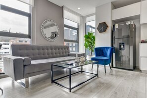 A modern living room with a grey sofa, a glass coffee table, a blue chair, a large mirror, a clock, and a refrigerator. - Area 93 Plus (Bogotá)