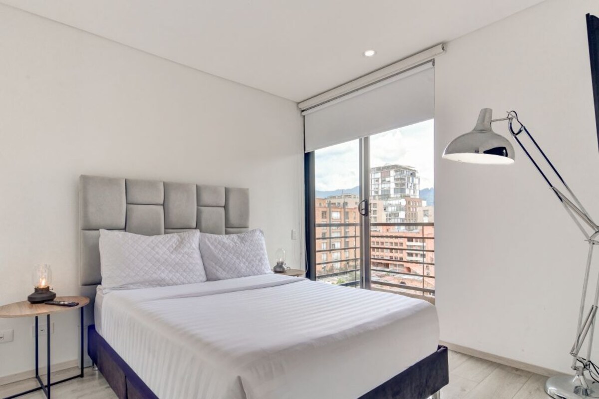 A modern bedroom with a large bed, a nightstand, a lamp, and a view of buildings through a window.