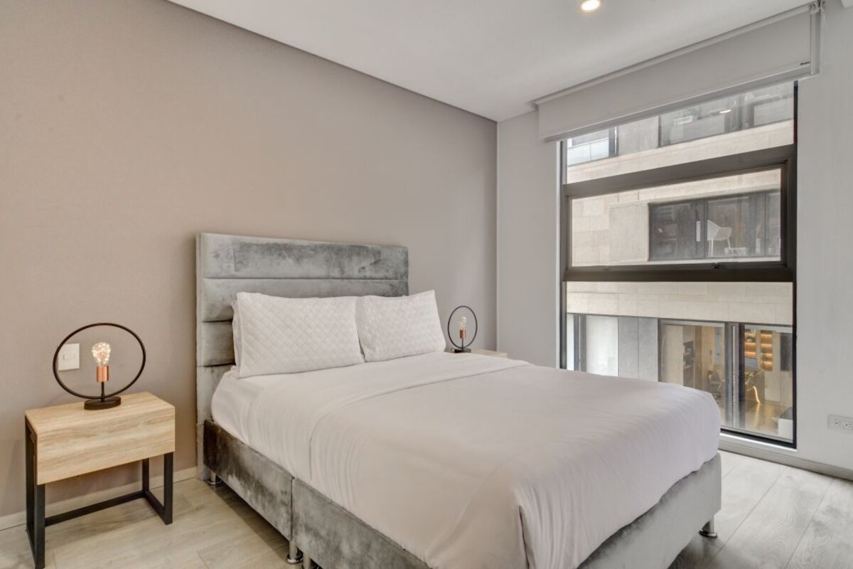 A modern bedroom with a large bed, a nightstand with a lamp, and a large window with a view of the city.