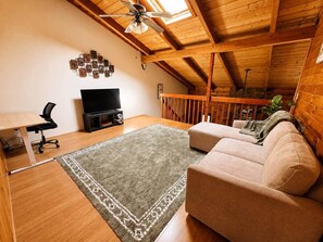 A loft-style room with a wooden ceiling, a flat-screen TV, a sofa, a desk, and a patterned rug. - Radford Retreat | 3 Min to RDU | Games & Arcade (Radford)