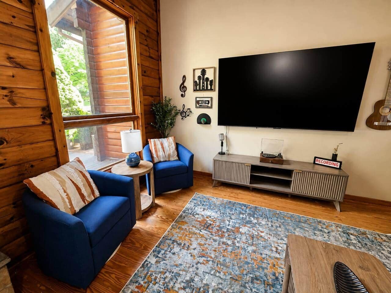 A living room with a large TV, two blue sofas, a patterned rug, and a wooden cabinet with a record player.