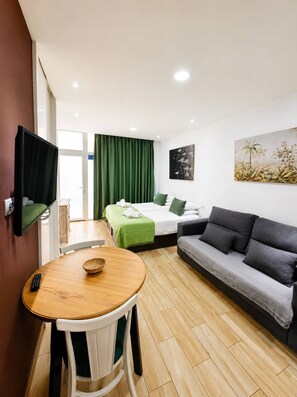 A modern hotel room with a wooden dining table, a sofa, a bed with green bedding, a television mounted on the wall, and a painting above the sofa. - 404|Apartment with ocean views|Canteras Beach (Las Palmas de Gran Canaria)