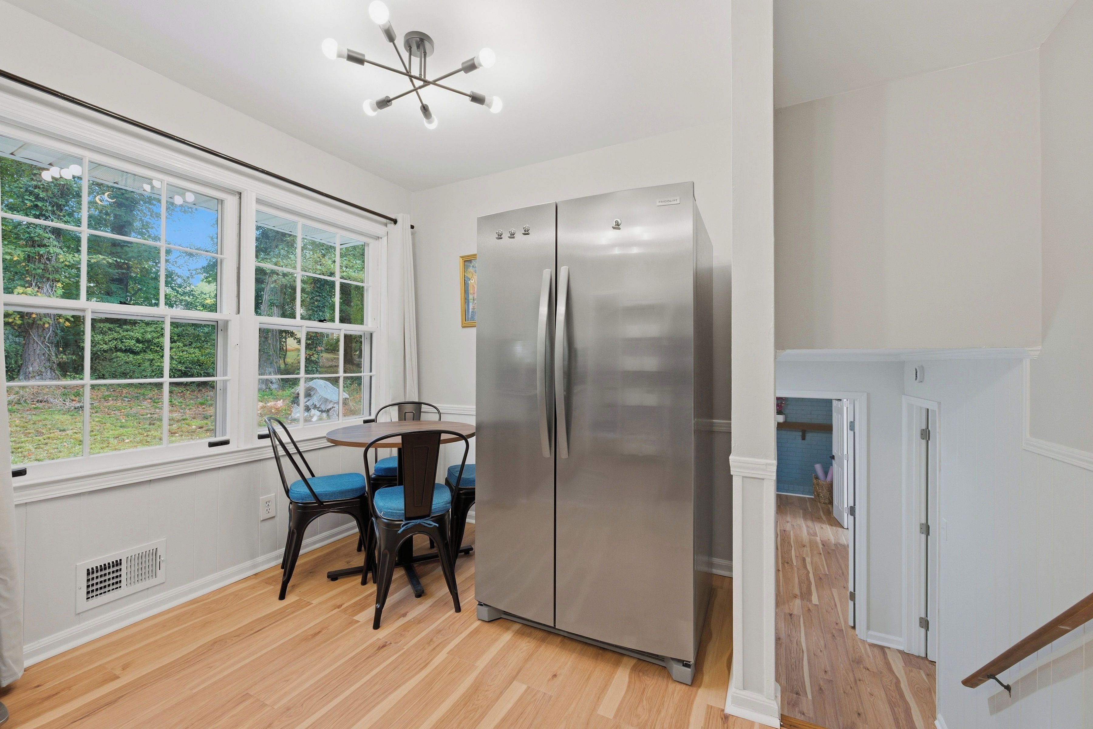 A kitchen with a stainless steel refrigerator, a dining table with chairs, and a window with a view of greenery.