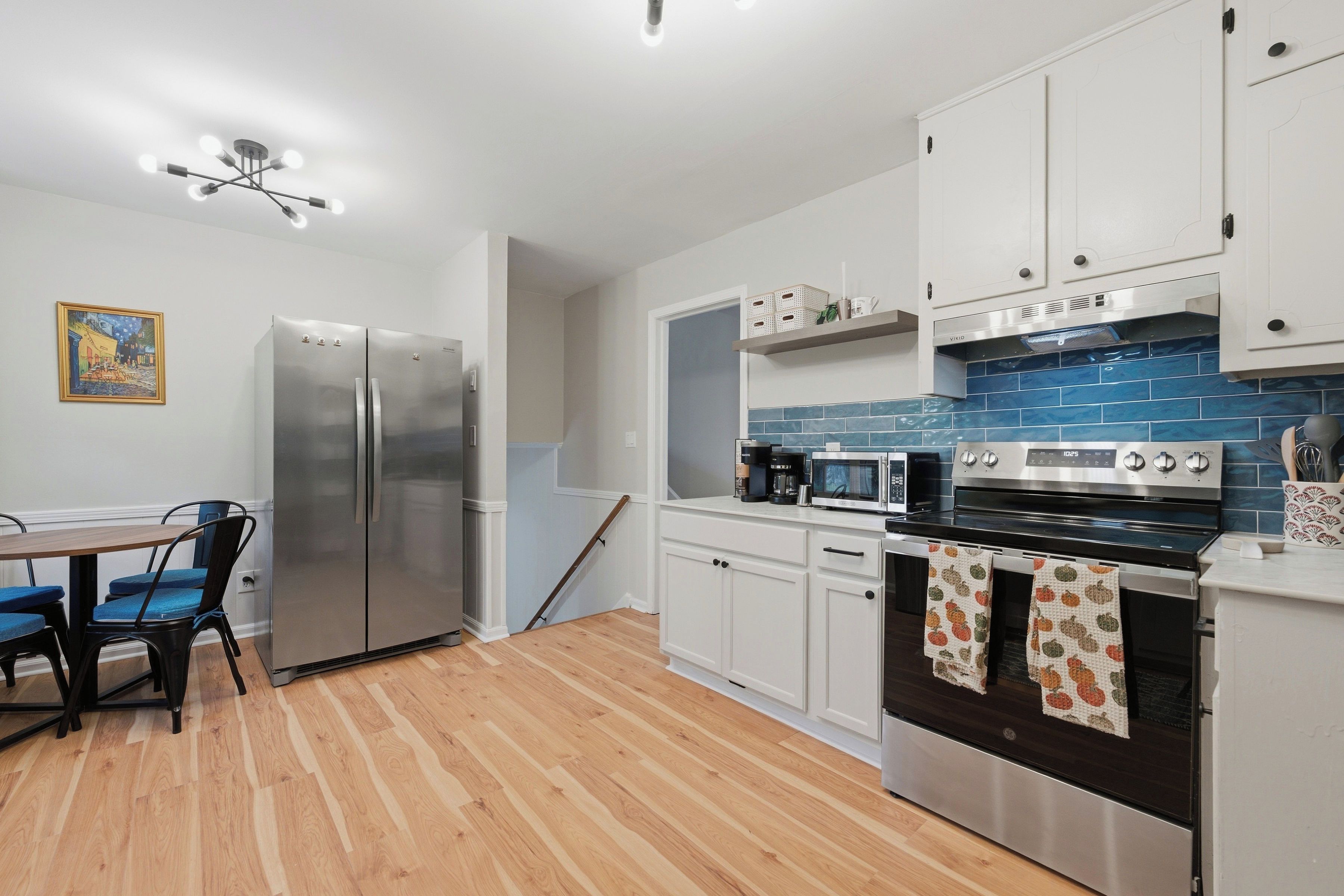 A modern kitchen with a stainless steel refrigerator, a microwave, an oven, and a coffee maker. There is a dining table with chairs and a painting on the wall.
