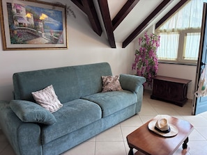 Ein blaugrünes Sofa mit gemusterten Kissen, ein Couchtisch aus Holz mit einem Hut, ein Gemälde einer Küstenlandschaft und ein Fenster mit Vorhängen.