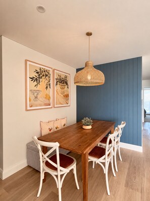 Ein Essbereich mit einem Holztisch, weißen Stühlen, einer gewobenen Pendelleuchte und zwei gerahmten Pflanzenbildern an der Wand.