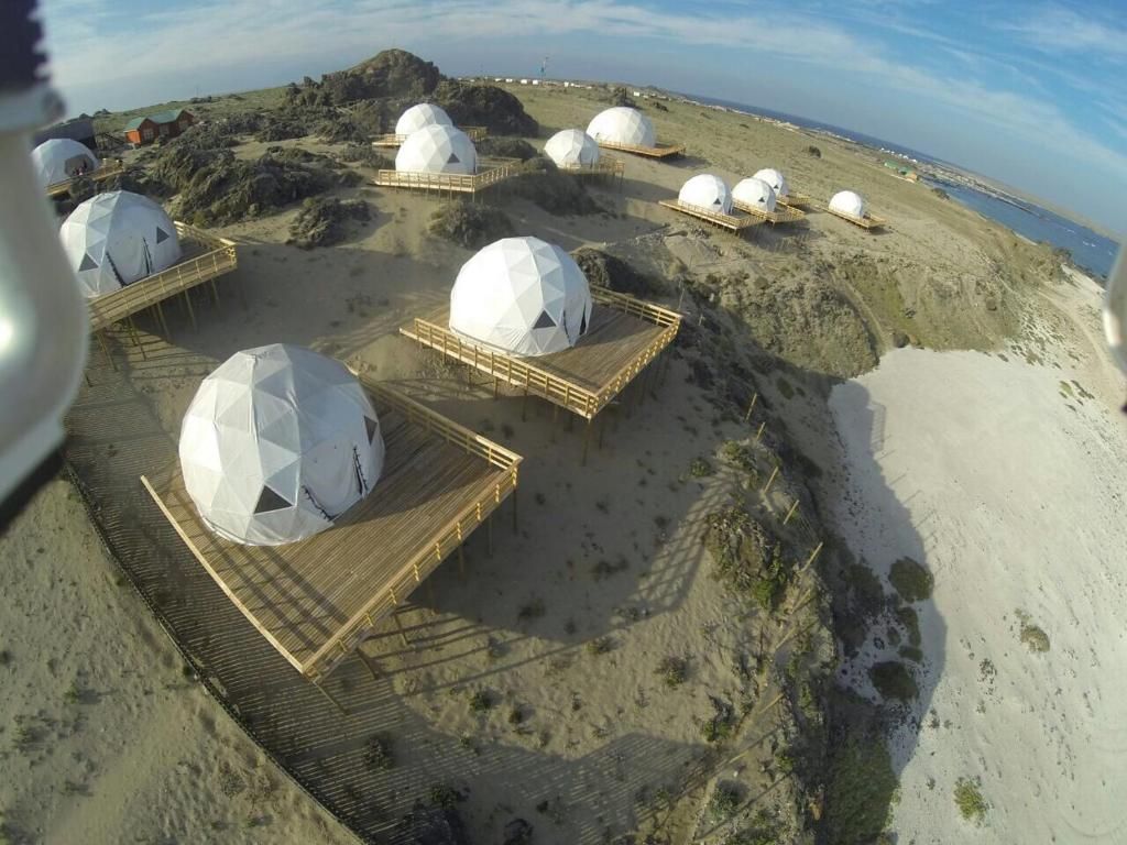 A series of dome-shaped structures with wooden walkways, set on a sandy terrain with a view of the sea.