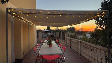 Un patio exterior con mesa y sillas, una pérgola con luces colgantes y vistas al atardecer.