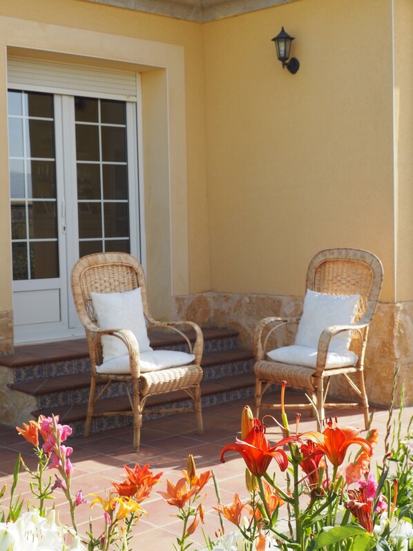 Two wicker chairs with white cushions, a tiled patio, and a yellow wall with a window and a wall-mounted light fixture.