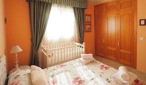 A bedroom with a crib, a bed with floral bedding, a nightstand, a lamp, and a wardrobe.