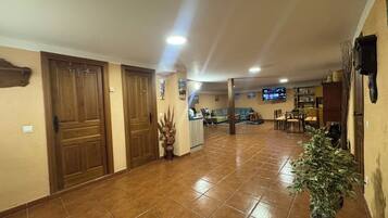 Un pasillo amplio con piso de baldosas, puertas de madera, un televisor montado en la pared y una zona de comedor con sillas y una mesa.