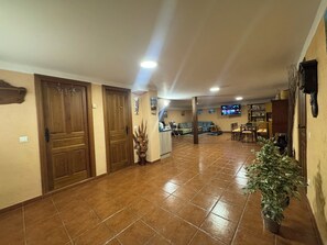 Un hall d’entrée spacieux avec un sol carrelé, des portes en bois, un téléviseur fixé au mur et un coin repas comprenant des chaises et une table.
