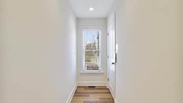 Un estrecho pasillo con suelo de madera, paredes blancas y una ventana con persianas.