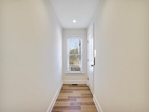 A narrow hallway with a wooden floor, white walls, and a window with blinds. - Beachside Luxury | Spacious 7BR + Porch + Parking! (Bethany Beach)