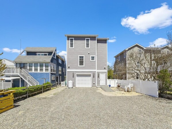 A two-story residential building with a garage, a covered driveway, and a small fenced area with outdoor furniture. - Beachside Luxury | Spacious 7BR + Porch + Parking! (Bethany Beach)
