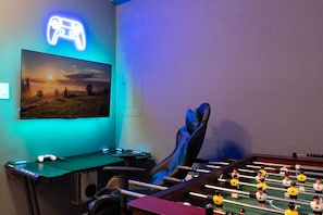 A gaming room with a neon game controller sign, a mounted TV, a foosball table, and a gaming chair. - Chic 6BR, Pool, Themed Rooms, Near Disney (Kissimmee)