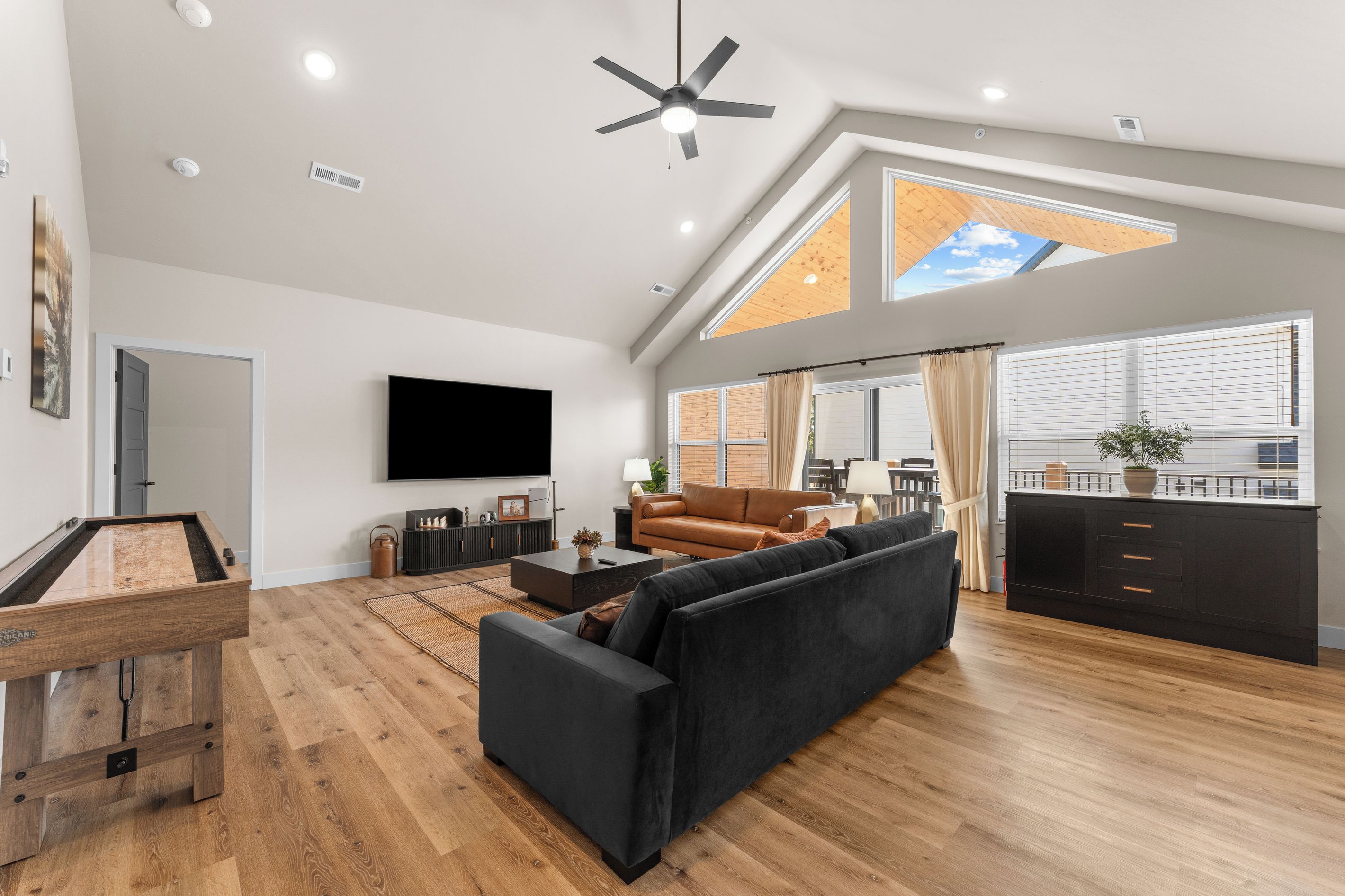 Ein geräumiges Wohnzimmer mit einer schwarzen Couch, Holzböden, einem Flachbildfernseher und einem Deckenventilator.
